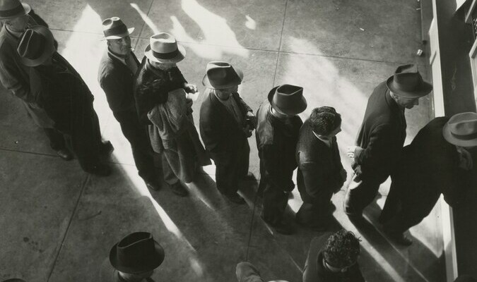 1938 - Unemployment benefits aid begins. Line of men inside a division office of the State Employment Service office at San Francisco, California, waiting to register for benefits on one of the first days the office was open. Photographer - Dorothea Lange